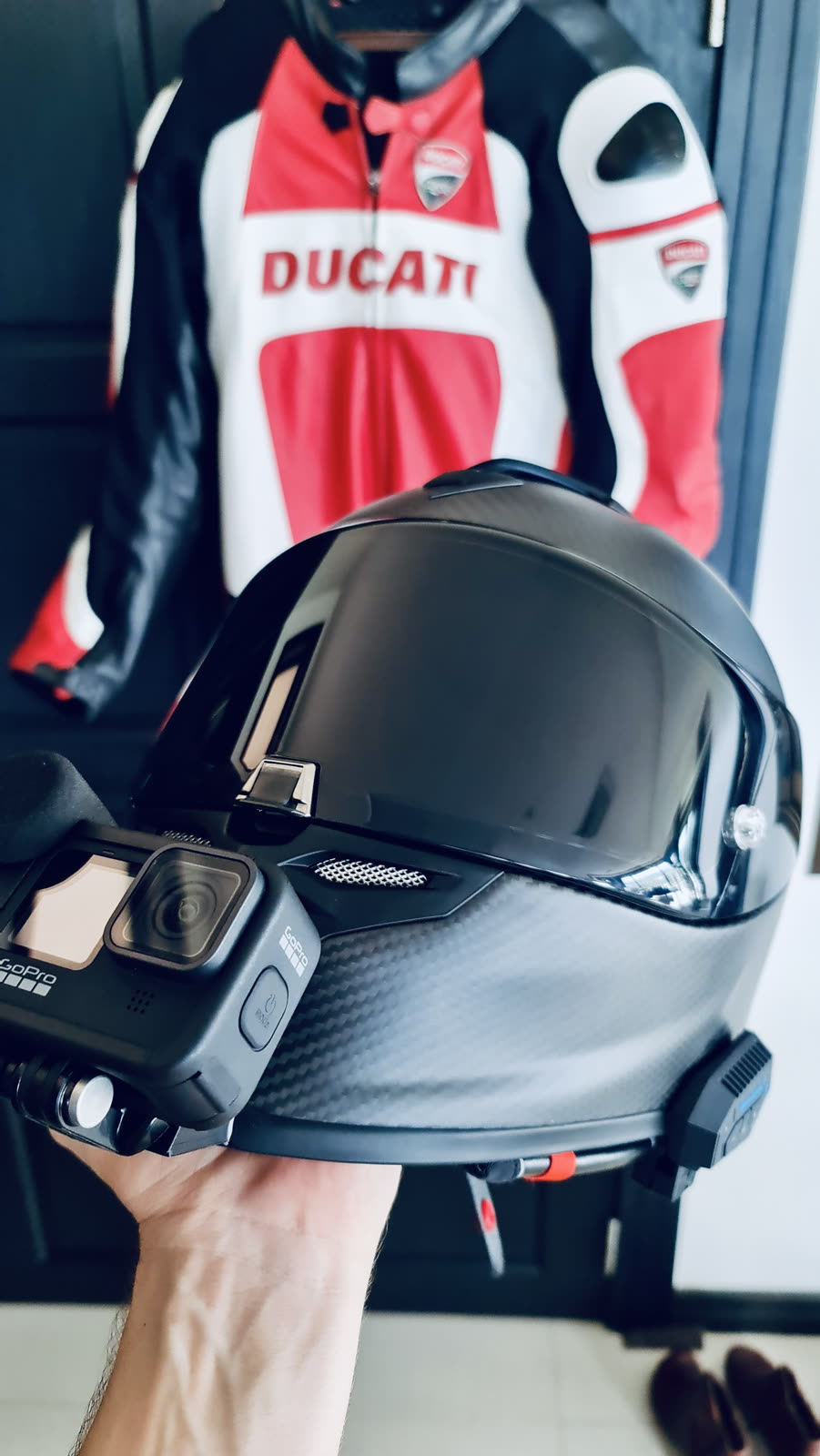 hand holding a GoPro camera beside a motorcycle helmet with a Ducati racing jacket in the background